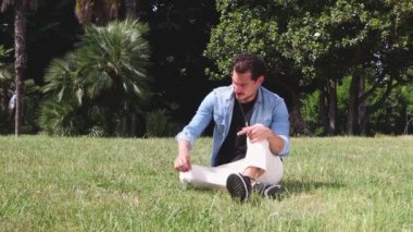 Mavi gözlü genç adam kamu parkında çimlerin üzerinde oturuyor. Adam kot gömlek ve bej pantolon giyiyor..