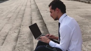 Genç iş adamı dışarıda bilgisayarıyla çalışıyor. Adam ofise gitmeden önce merdivenlerde oturuyor..