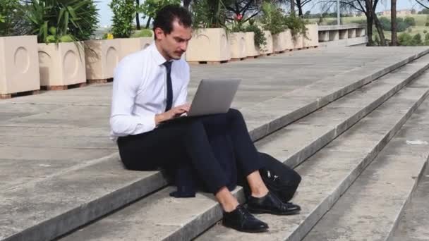 年轻的商人正在外面用他的电脑工作 这个人在去办公室之前先坐在楼梯上 — 图库视频影像