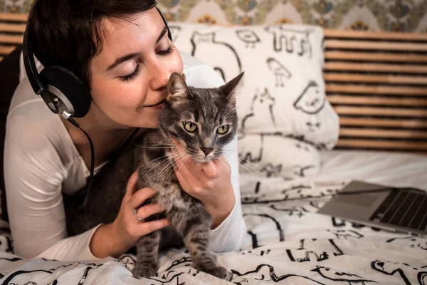 Beautiful woman plays with the cat on the bed. The young woman listens to music on headphones while she is petting her cat.