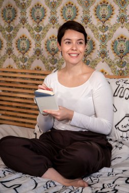 Woman is writing on her personal organizer. The beautiful woman is sitting on the bed and smiling thinking about what she has to write.