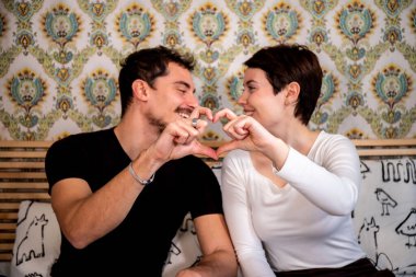 Young couple is sitting on the bed and looking into each other's eyes. The couple forms a heart with their hands. Concept of love and happiness.