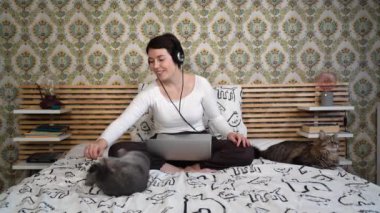 Beautiful woman is working on the computer while listening to music on headphones. The woman is sitting on the bed and petting her cat.
