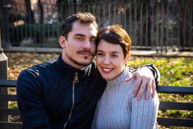 Young couple is sitting on a park bench in Rome. The man is looking towards the camera while the damage is laughing..