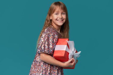 Parlak, zarif elbiseli sevimli kız çocuğu elinde turkuaz bir arka planda kırmızı bir hediye kutusu tutuyor.