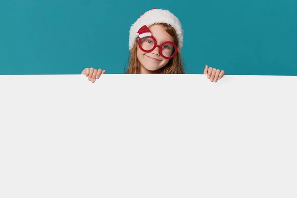 サンタクロースの帽子のかわいい女の子とターコイズの背景のパーティーグラス 子供は広告のための記号 テキストのための空のスペースを保持します うわ面白い顔 — ストック写真