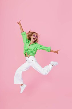 Sıradan giyinmiş, pembe bir arka planda zıplayan ve kutlama yapan neşeli genç bir kadının tam boy portresi.
