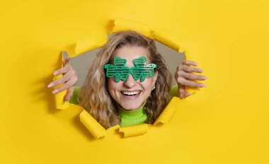 Surprised woman wearing green clover-shaped glasses looking, peeping through the bright yellow paper hole. Showing hand to side. Copy space for text. St Patrick day. Wow face 