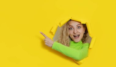 Surprised curly haired young lady have fun, poses in yellow paper hole for making photo, feels upbeat, expresses positive emotions. Wow face