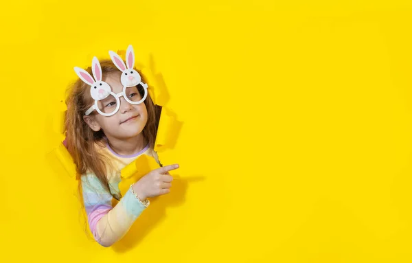 かわいいウサギの形のメガネを探しているかわいい驚きの子供の女の子は 黄色の紙の穴を覗きます イースター休暇のための子供用品を宣伝しています テキストのコピースペース — ストック写真
