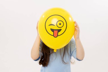 Çocuk bir kız, beyaz arka planda sarı göz kırpan bir ifadenin arkasına saklanıyor. Komik surat. Dünya Emoji Günü