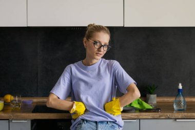 Portrait of  young tired tortured cleaning service woman. Housewife cleaner in yellow rubber gloves stands in the kitchen after a general cleaning of the house