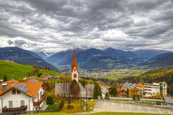İtalyan Dolomitlerinin arka planında bulutlu gökyüzü olan kilise ve turuncu renkli çatıları olan bir köy.