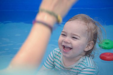 Sıcak bir yaz gününde annesinin yanında yüzme havuzunda oturan gülümseyen sarışın çocuk. Anne ve oğul birlikte serinlemek ve oynamak için suyun tadını çıkarırken neşe ve eğlence anları paylaştılar.
