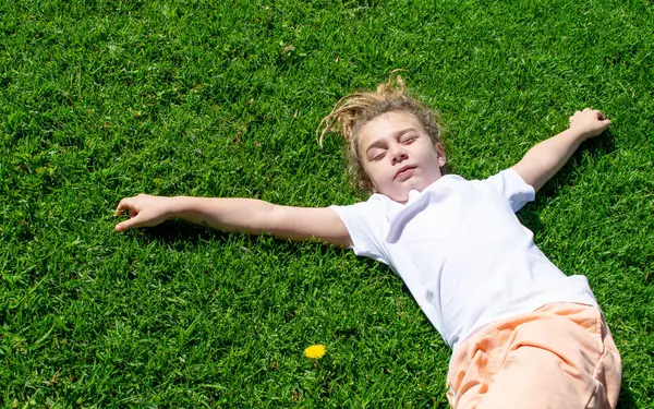 A blonde boy, dressed for summer, relaxes in the sunny field. He enjoys the day outdoors, lying on the grass while the sun\'s rays illuminate his golden hair, creating a moment of happiness and serenity.