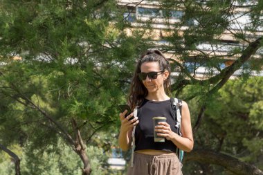 Genç ve güzel bir kadın sokakta çimenlerde otururken cep telefonu kullanıyor ve kahve içiyor..