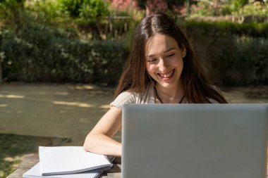 Üniversite öğrencisi, okulun başında dizüstü bilgisayarını kullanırken gülümsüyor. Teknoloji, akademik hayat, sanal toplantı, üniversite diploması