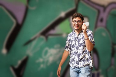 Güneş gözlüklü modern genç adam şehirde kredi kartını gösteriyor. Şehir modası, finans, yaşam tarzı.