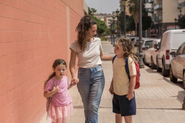 Genç anne güneşli bir günde çocuklarını sırt çantasıyla okula götürür. Okula geri dönmek, okula dönmek, okulun ilk günü, aile, gülümseme, okula gitmek.