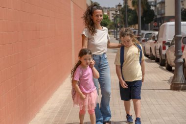 Genç anne okulun ilk gününde çocuklarıyla birlikte okula yürür. Güneşli bir günde konuşur ve gülümserler. Okula dönüyorum, mutlu bir aile, sırt çantaları, okula gidiyorum..