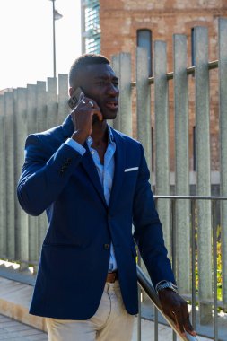 Gülümseyen Afrikalı Amerikalı yönetici iş alanındaki akıllı telefonuyla işe yürüyor, gökdelenler ve ofis binaları arasında.