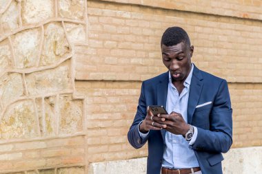 Şaşkın bakışlı zarif takım elbiseli genç siyah yönetici, yürürken akıllı telefon kullanıyor. Modern iş ve teknoloji kavramı