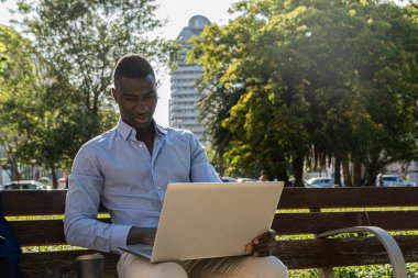 Yoğunlaşmış Afro-Amerikan yönetici, dışarıda laptopu ve bir fincan kahveyle çalışıyor. Uzak çalışma, iş ve doğal ortamın tadını çıkarma kavramı.