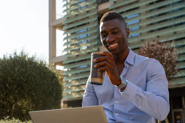 Gülümseyen genç Afro-Amerikan iş adamı dizüstü bilgisayarı ve kahvesiyle finans bölgesinde çalışıyor iş dünyası, teknoloji, uzaktan çalışma, şehir.