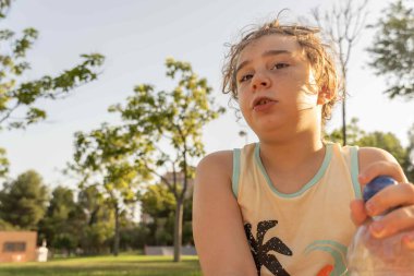 Açık renk saçlı çocuğun yakın çekimi, el kol hareketi yaparken elinde bir şişe su tutuyor, parkta oturuyor. Güneşli sıcak bir gün. Doğal arkaplanda metin için boşluk.