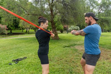 Bir adam ve bir kadın, halka açık bir parkta egzersiz yapmak için direniş bantlarını kullanıyor. Açık alandaki güç ve forma odaklanıyor..