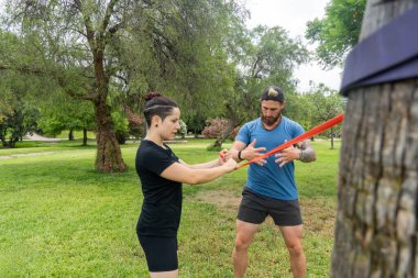 Yeşillik bir parkta direniş bantları kullanarak dayanıklılık eğitimi alan iki birey, direniş eğitimine odaklanmış açık hava fitness rutinine girişiyor.