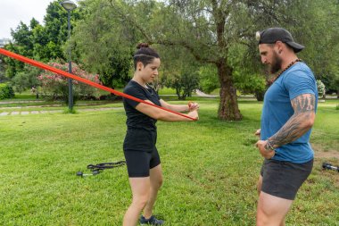 Bir direniş bandı antremanına katılan iki kişi parkta egzersiz yaparken görülüyor. Açık hava fitness ve güç antrenmanı aktivitelerini vurguluyorlar..