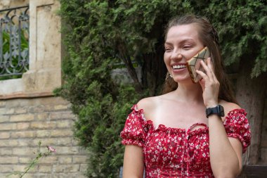 Kırmızı çiçekli elbiseli neşeli kızıl saçlı bir kadın, gün ışığında telefonunda mutlu bir şekilde sohbet ederken görülüyor..