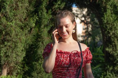 Kırmızı çiçekli elbiseli neşeli kızıl saçlı bir kadın, gün ışığında telefonunda mutlu bir şekilde sohbet ederken görülüyor..