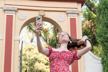 Kırmızı çiçekli elbiseli neşeli kızıl saçlı bir kadın, gün ışığında telefonunda mutlu bir şekilde sohbet ederken görülüyor..