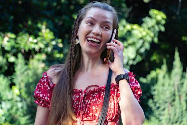 Kırmızı çiçekli elbiseli neşeli kızıl saçlı bir kadın, gün ışığında telefonunda mutlu bir şekilde sohbet ederken görülüyor..