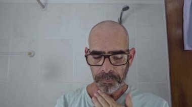 Ev banyosunda gözlüklü kel bir adam sakalını pille çalışan bir tıraş makinesiyle tıraş etmeye başlar. Günlük kişisel bakım rutini ev ortamında yakalanır..