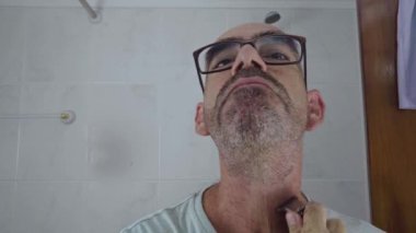 Gözlüklü, kel ve yetişkin bir adam sakalını tıraş ediyor. Ev banyosunda pille çalışan bir jilet kullanıyor. Sıradan bir erkek günlük rahatlama ortamında tımar ediliyor..