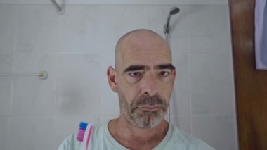 Yetişkin, kel bir adam banyoda dişlerini fırçalarken aynaya neşeli bir ifadeyle bakıyor ve diş bakımı rutininden tatmin oluyor..
