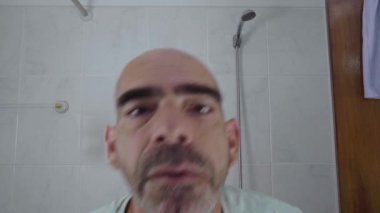 Yetişkin ve kel bir adam diş fırçalama rutinini ev banyosunda tamamlar. Aynada temiz dişlerini zevkle gözlemler. Temiz gülümsemesi ve hijyeniyle tatmin olur..