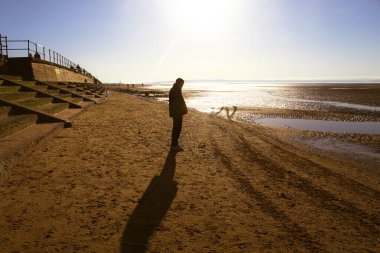 Sahilde uzun bir yürüyüş yapan bir erkeğin çarpıcı manzarası. Parlak güneşe doğru yürüyor. Gölgesi kumda açıkça görülüyor. Bu egzersiz hem fiziksel hem de zihinsel sağlık için iyidir..