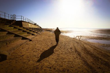 Sahilde uzun bir yürüyüş yapan bir erkeğin çarpıcı manzarası. Parlak güneşe doğru yürüyor. Gölgesi kumda açıkça görülüyor. Bu egzersiz hem fiziksel hem de zihinsel sağlık için iyidir..