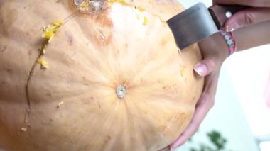 Cadılar Bayramı partisi için büyük turuncu balkabağı oyan dişi, evdeki ahşap masada otururken, şaka mı şeker mi havasında olmak için bıçağıyla balkabağı üzerinde korkunç bir yüz ifadesi yapıyor.