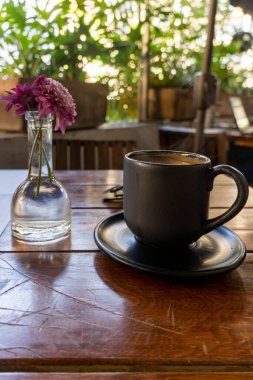 Siyah killi bir fincan kahve, ahşap masa, arka planda bitki örtüsü, ve masayı süsleyen çiçekler, Meksika Latin Amerika