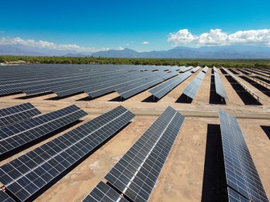Kısmen bulutlu bir gökyüzünün altında geniş güneş panelleri Tinogasta 'da uzanır ve Andes Dağları' nın silueti uzaktan görünür. Arjantin 'in alternatif enerjilere doğru ilerlemesinin bir sembolü..