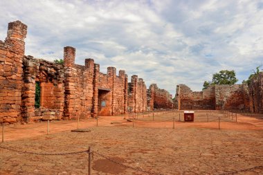 San Ignacio Mini, Misiones, Arjantin harabelerinin iç manzarası. Çatısız taş duvarlar, antik yapı ve arka planda sunağın kalıntıları. Taş zemin, kısmen bulutlu gökyüzü ve çevreleyen bitki örtüsü.