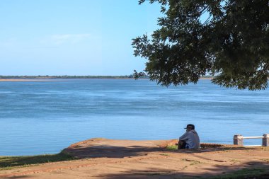 Paso de la Patria 'nın rıhtımında oturan genç bir adam, Arjantin, Corrientes, Parana Nehri' ne melankolik bir şekilde bakıyor. Yolun karşısında, Paraguay kıyısı bir ağacın altında, su ve kara bir yansıma atmosferinde.