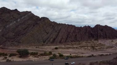 Fiambala, Catamarca 'daki Caadon del Indio' ya yaklaşan çarpıcı hava görüntüsü. Dağlar, geçitler, neredeyse kuru nehir, bulutlu gökyüzü, 6 millik yol, ve görüş açısına göre araçlar görünür..