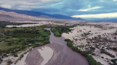 Medanitos yakınlarındaki Fiambala ya da Saujil Nehri 'ni gösteren bir video. Arka planda, yaklaşan bir fırtına ve gri gökyüzünün altında, çöl manzarasını ve kum tepelerini aydınlatan görkemli And Dağları var.