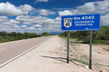 Arjantin 'in Katamarca şehrine doğru Km 4040' ı işaret eden ikonik 40. yolda, parçalı bulutlu bir gökyüzünün altında, mavi bir yol işareti var.